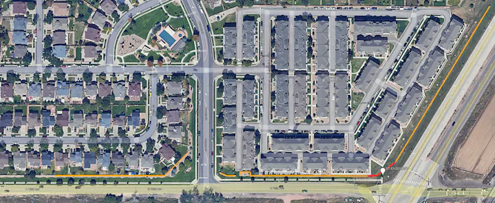 Aerial view of a suburban neighborhood featuring houses, streets, and a park area, with a nearby highway and pathways.