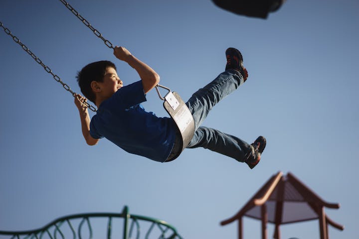A boy joyfully swings on a swing set against a clear blue sky, enjoying his time at a playground.