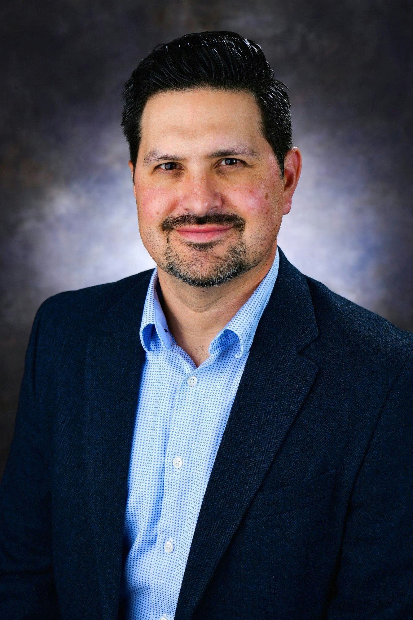 A person in a suit and shirt smiles against a neutral backdrop.