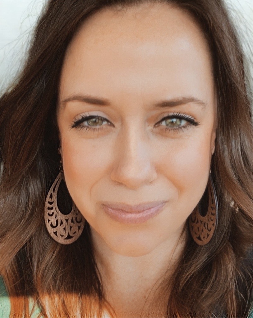 A close-up portrait of a woman with long hair, wearing decorative earrings and a subtle smile. Natural lighting enhances her features.