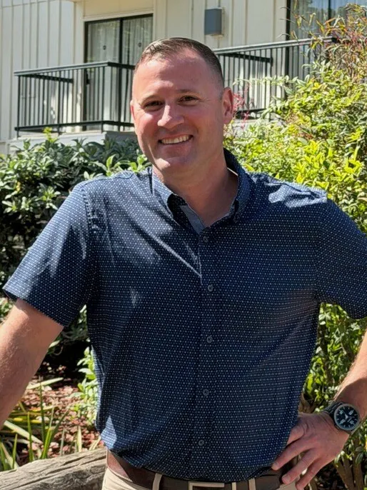 A smiling man stands by a wooden post in a landscaped area with plants and a building in the background. He wears a short-sleeved shirt.