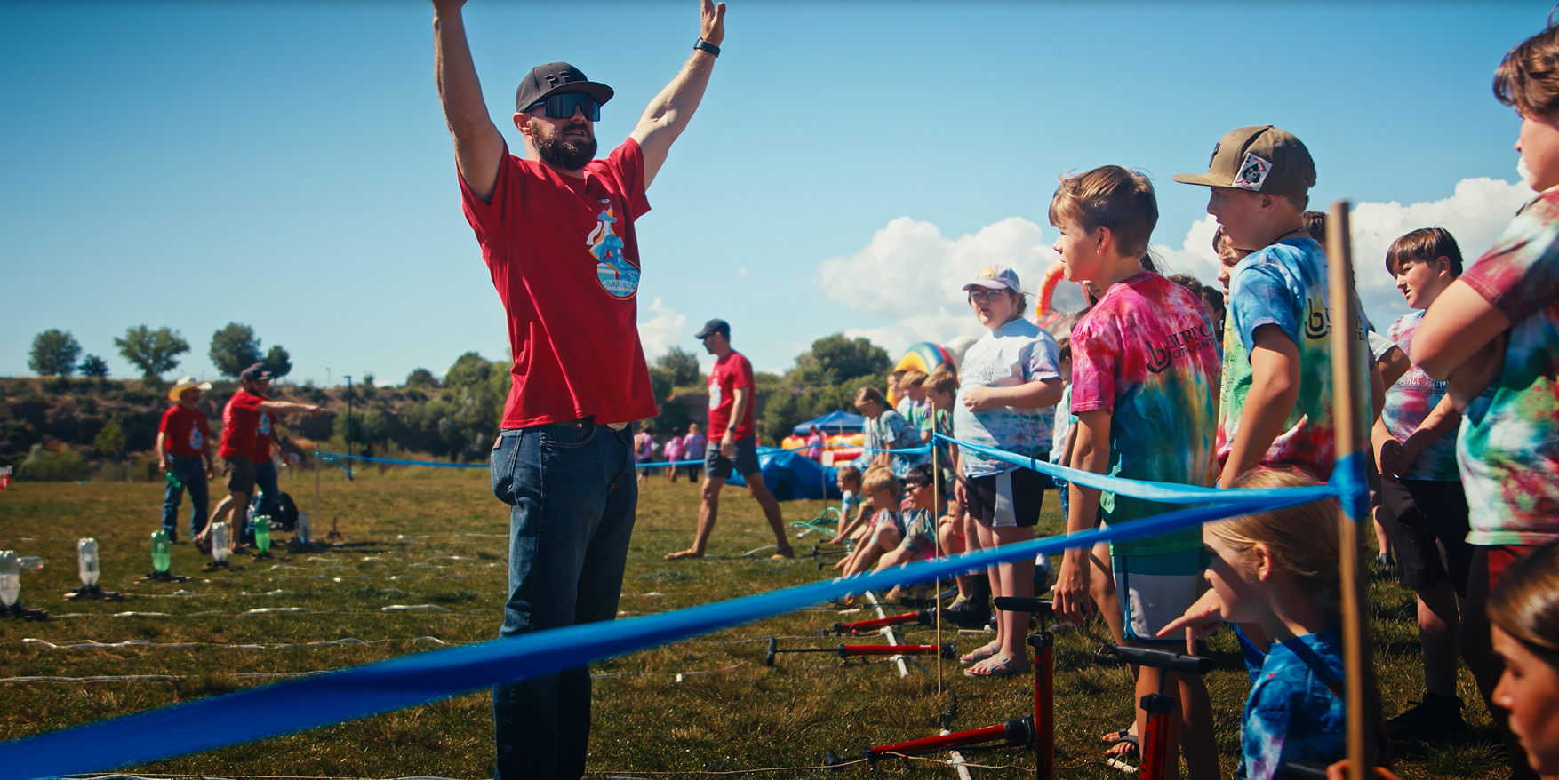 A man in a red shirt raises his arms, engaging with children in a colorful outdoor event with blue barriers and grassy fields.