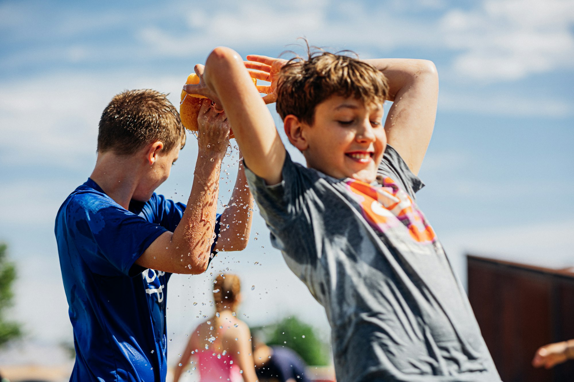 Two kids playing outside, splashing water with a sponge, smiling on a sunny day.