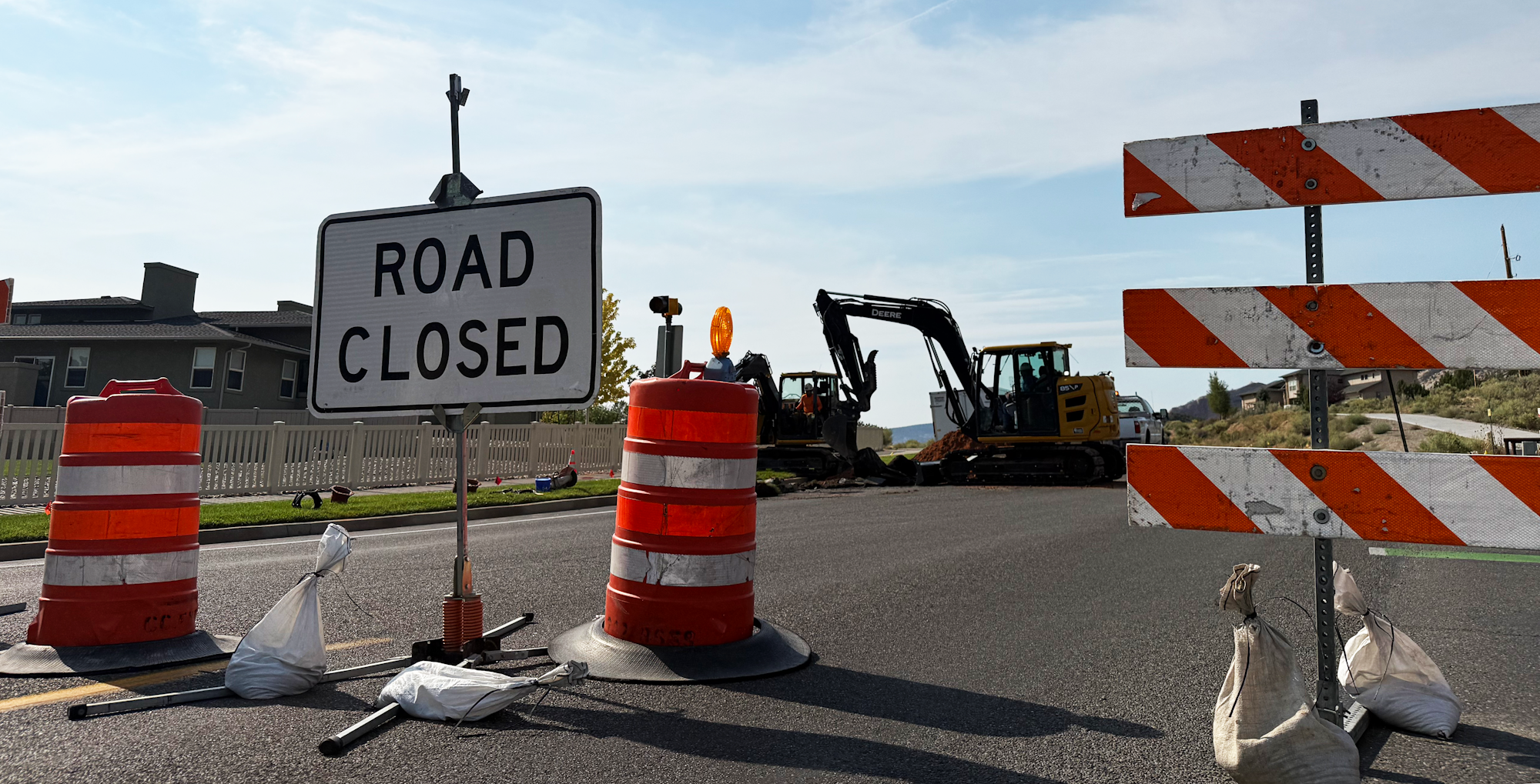 Road closed with construction barriers, cones, and excavators in the background under a clear sky.