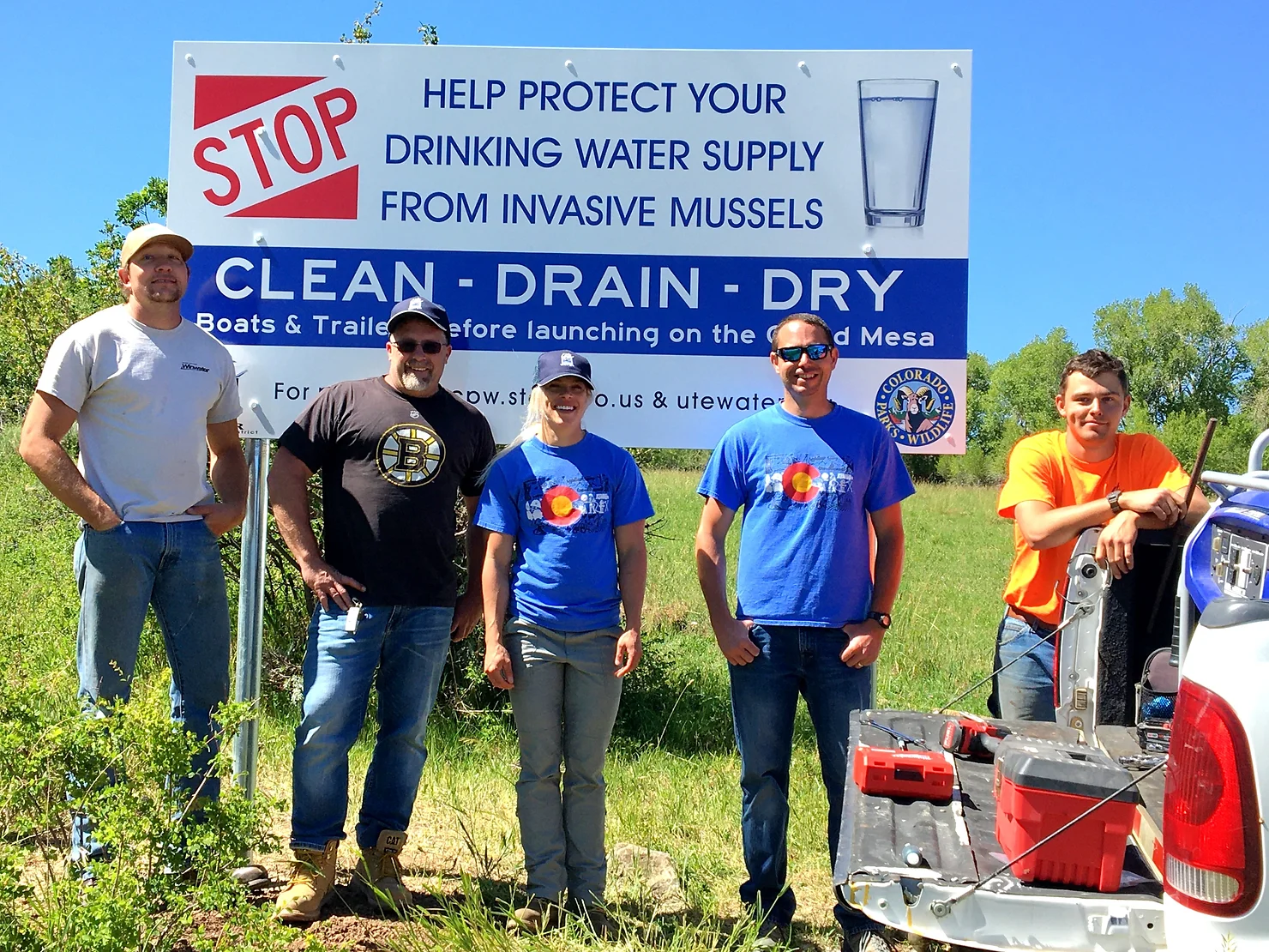 Five people posing by a sign promoting protection of water supply from invasive mussels by cleaning, draining, and drying boats and trailers.
