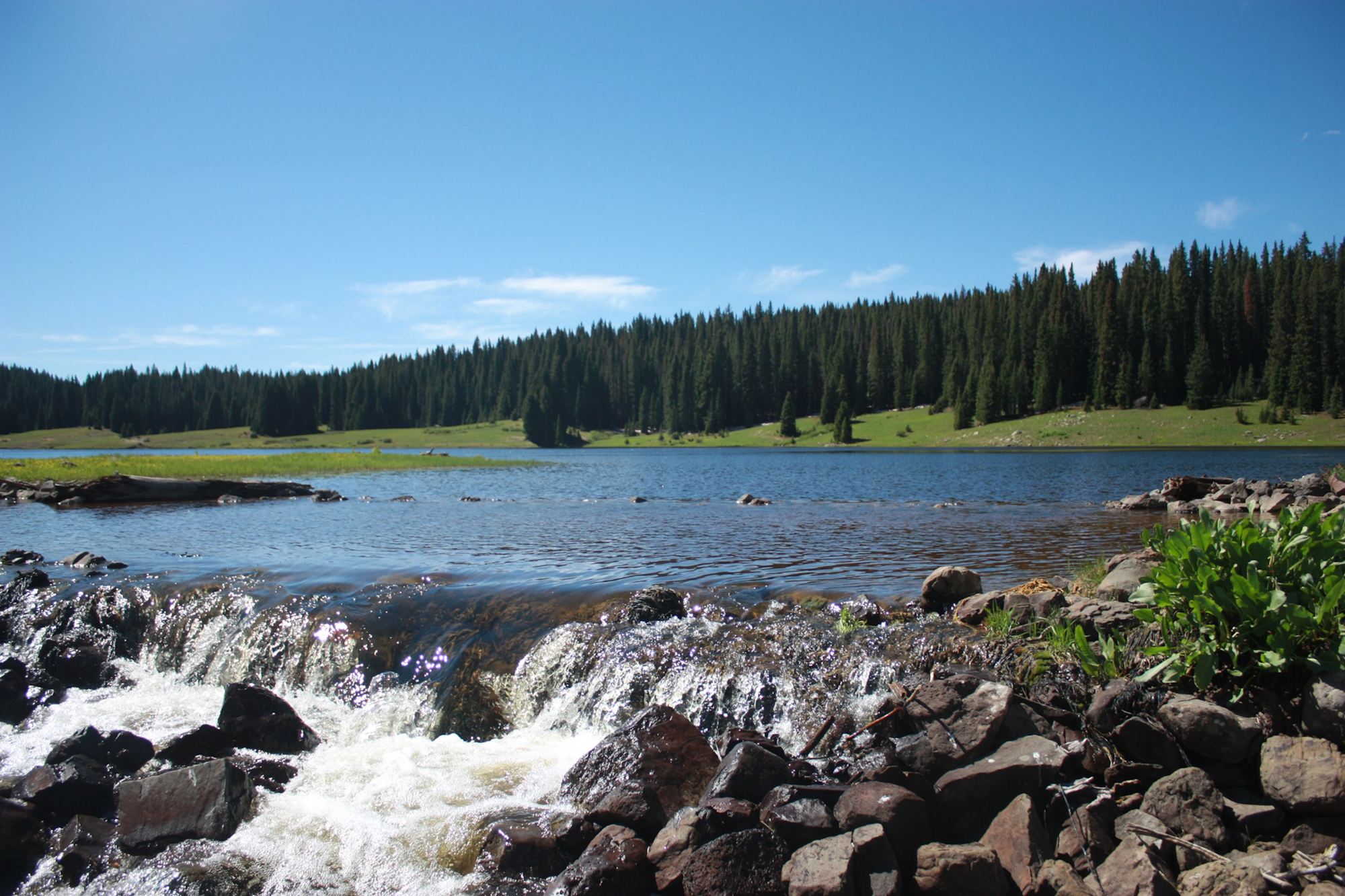A scenic mountain lake with a rocky stream in the foreground and pine trees under a blue sky.