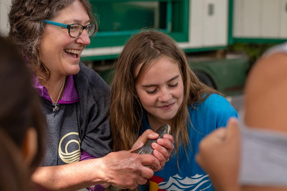 A smiling woman and a focused girl with a fish, learning in an outdoor setting.