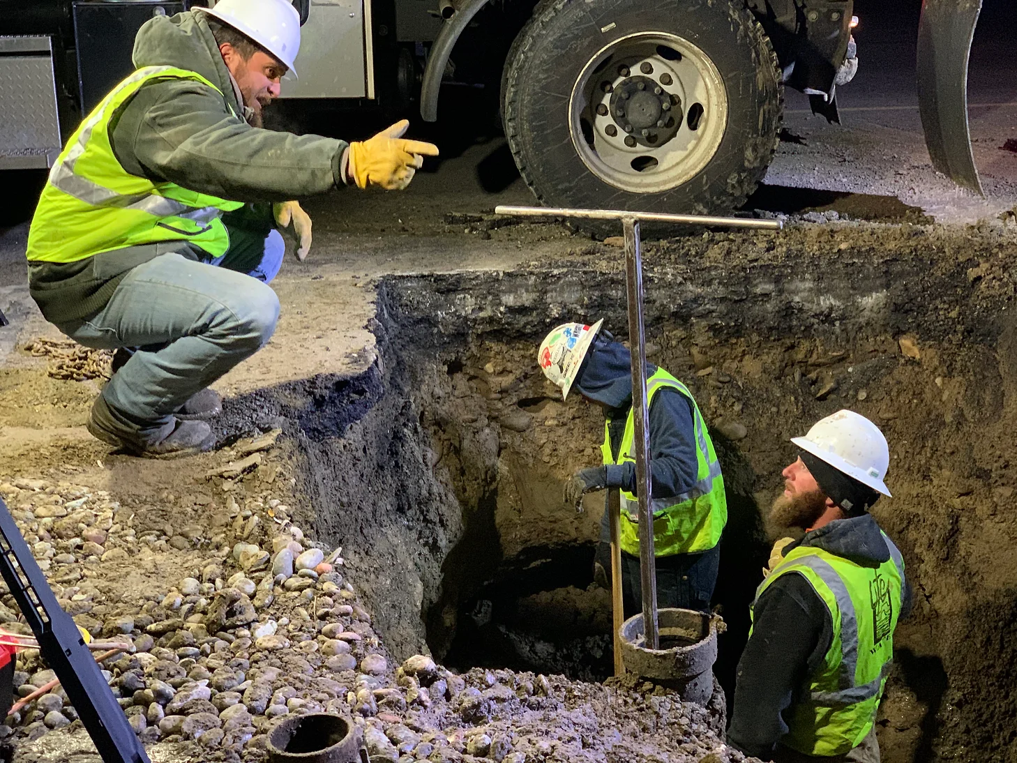 Three construction workers in high-visibility gear and helmets working at a nighttime excavation site.