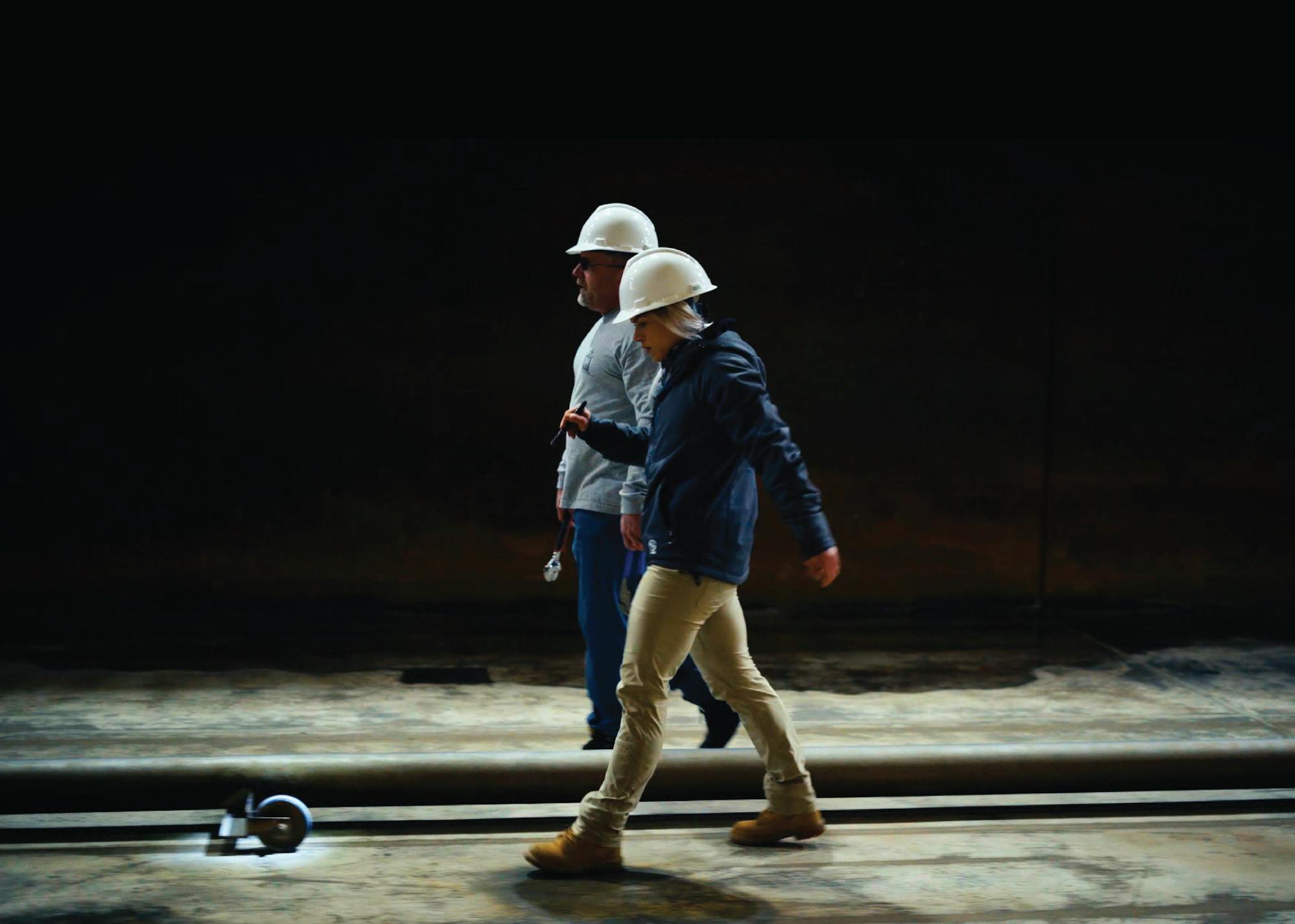 Two individuals in hard hats walk across a dimly lit industrial space, showcasing teamwork and focus on a task.
