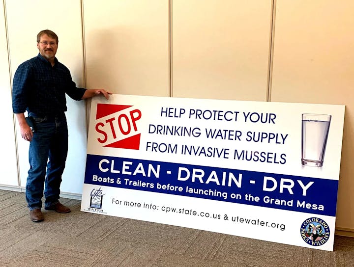 A man holding a sign promoting the protection of water supply from invasive mussels, advising to clean, drain, and dry boats and trailers.