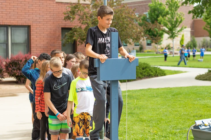 A group of kids watching a boy operating an outdoor electronic device at school.