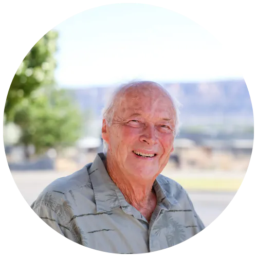 An elderly man smiles in a casual shirt, set against a scenic backdrop with trees and mountains.