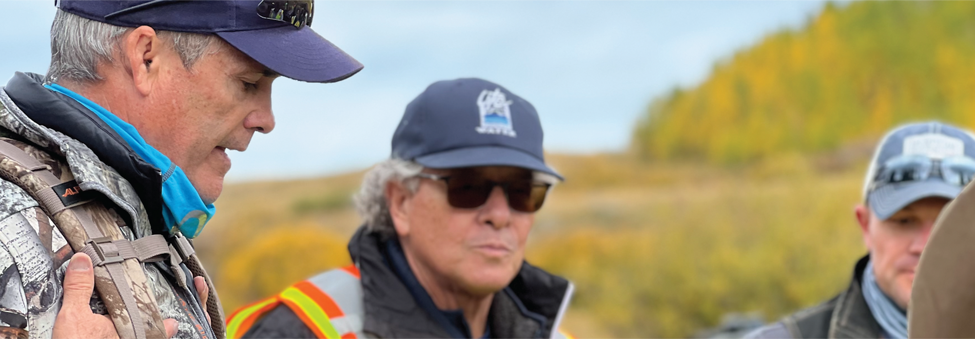 Three men with caps, one in a safety vest, outdoor setting with autumn trees.