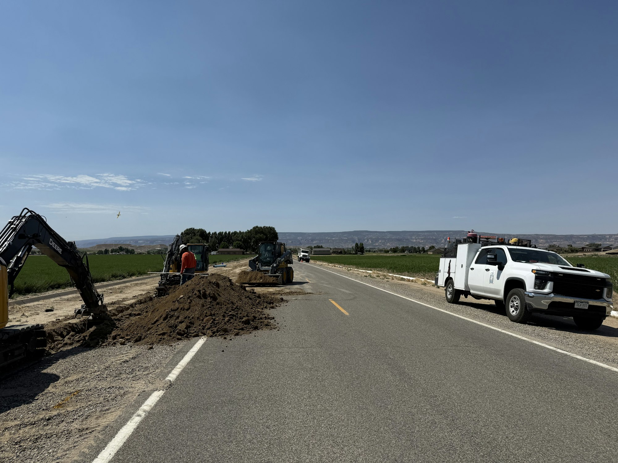 Roadwork scene with construction vehicles and workers beside a rural road. A white truck is parked on the right.