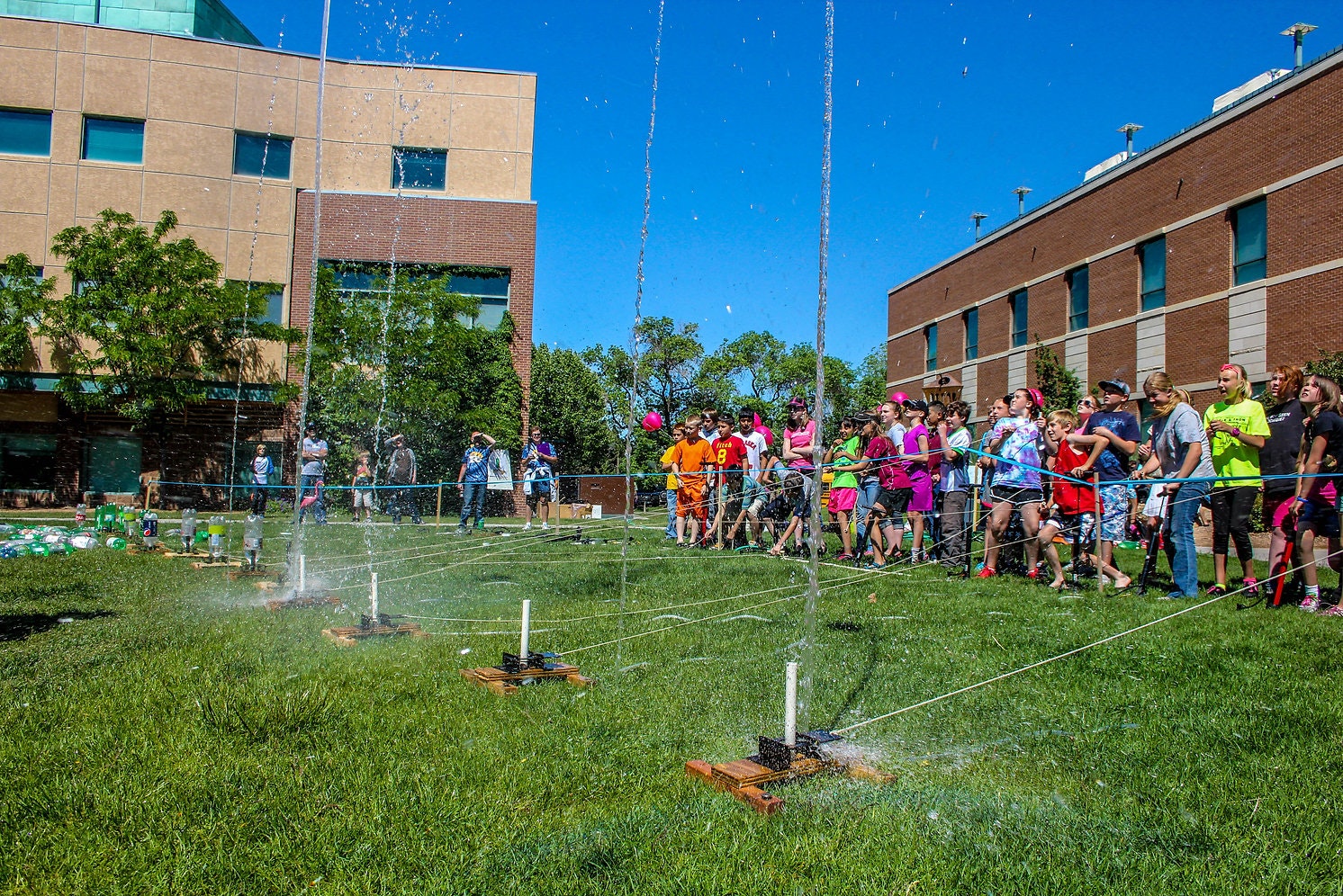 A group of people watching water rockets launch on a sunny day outdoors.