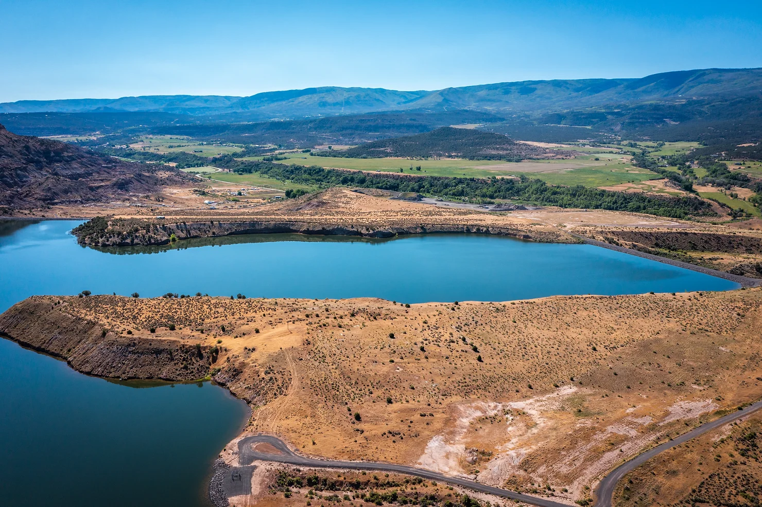 Aerial view of a winding lake with sparse vegetation surrounded by hills under a clear sky.