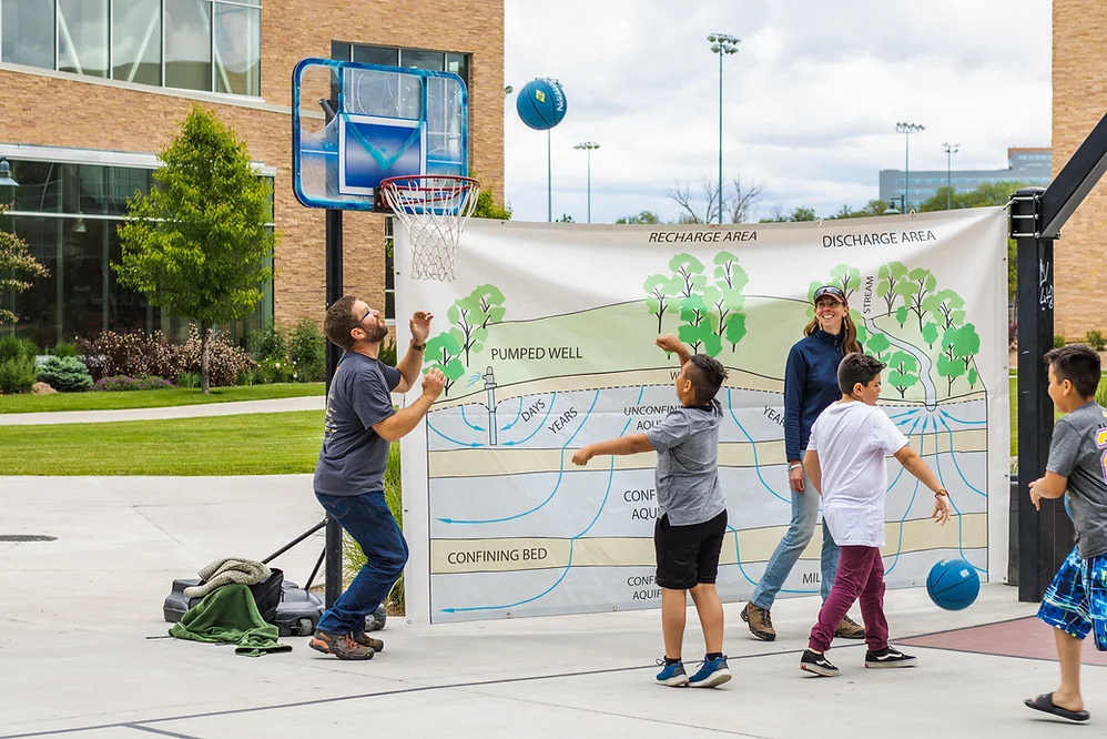 Adults and kids playing basketball with educational groundwater illustration in the background.