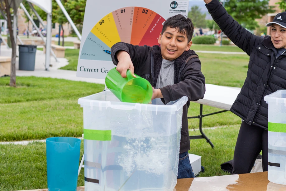 A boy pouring water from a pitcher into a clear bin with excitement as an adult cheers, at an outdoor event with educational materials.