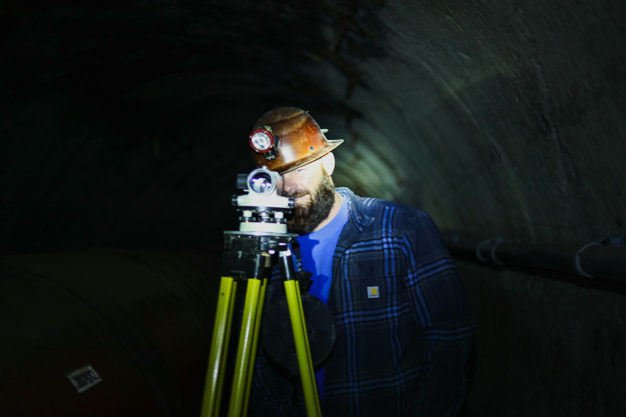 A man in dark tunnel with blue flannel, hardhat with light looking through lens
