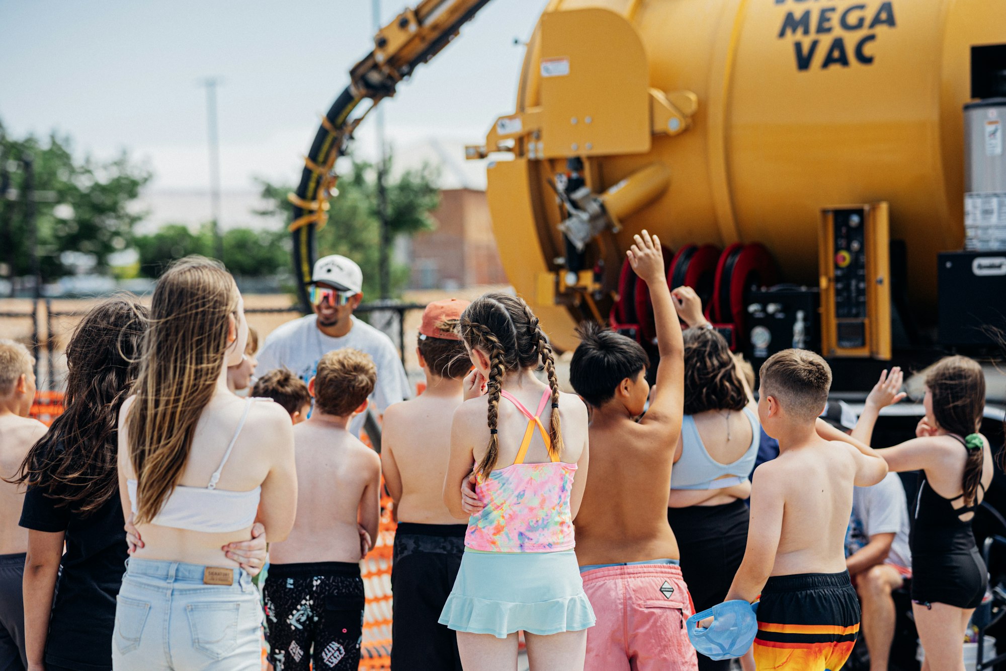 Group of kids gathered around a large yellow Mega Vac machine outdoors, some with raised hands.