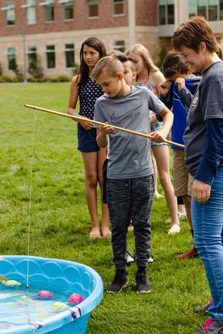 Children and an adult doing a fishing game with toy fish in a pool outdoors.