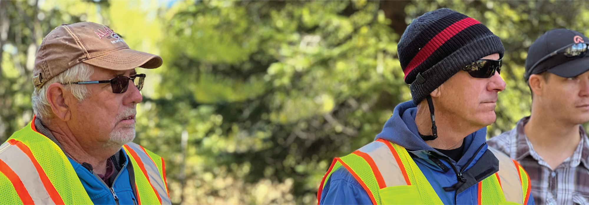 Three men in safety vests outdoors, one with sunglasses, another with beanie and sunglasses, and the third in a cap.