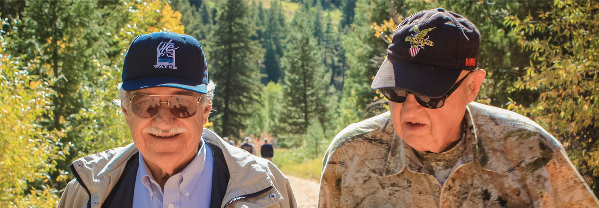 Two elderly men in baseball caps walking outdoors with trees in the background.