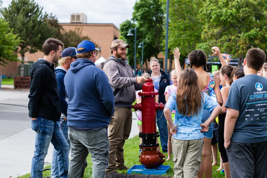 Group of people, including children, gathered around a fire hydrant outdoors, with a man gesturing happily.
