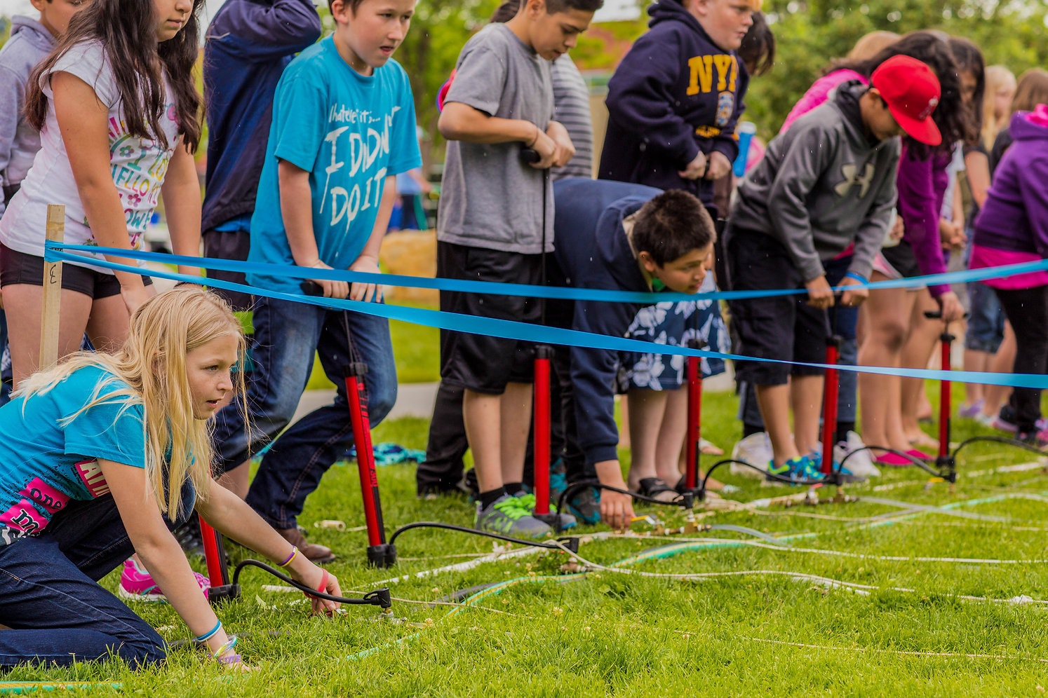 Children playing a water game outdoors, bending to manipulate hoses with water spray. Grass and colorful ribbons visible.