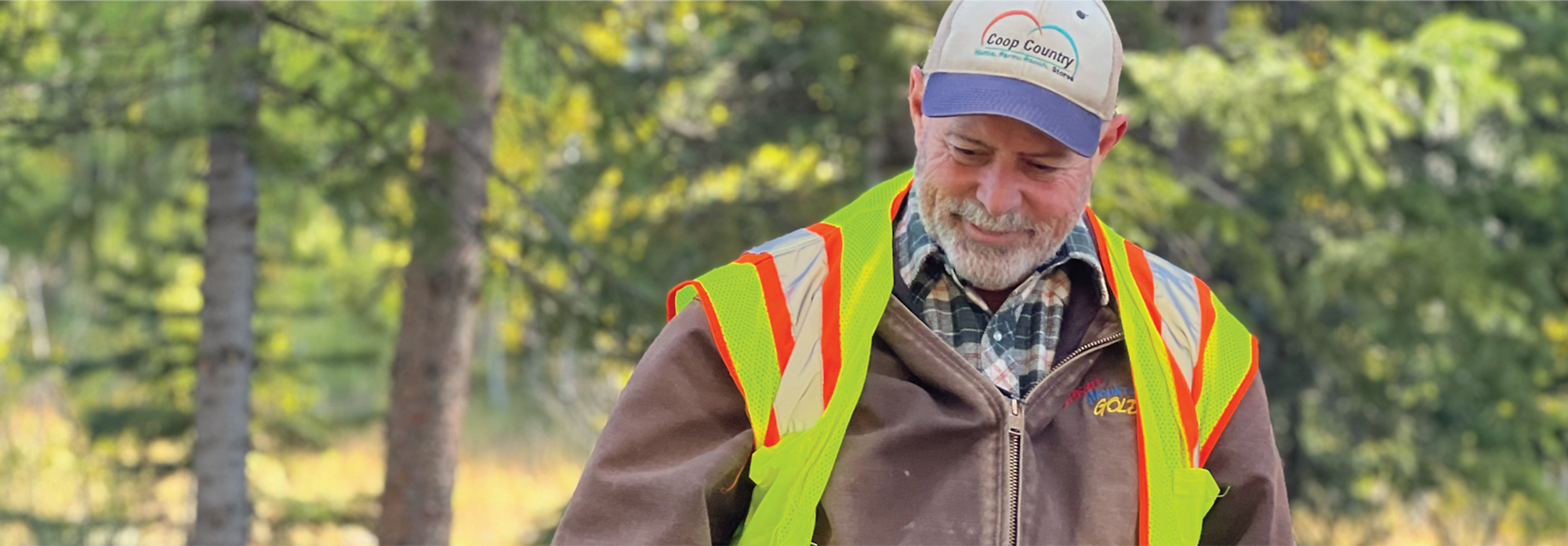 A smiling man in a high-visibility vest and baseball cap outdoors with trees in the background.