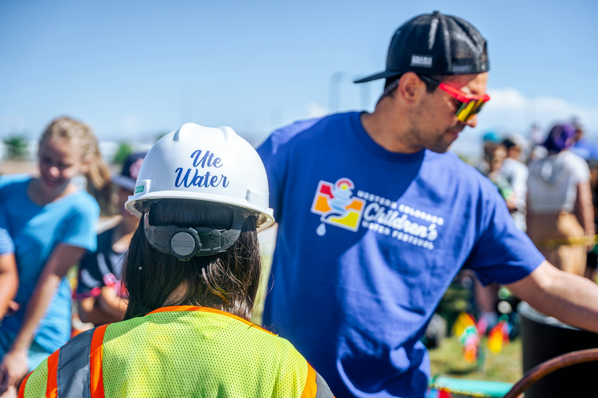 Person in safety gear with "Ute Water" helmet, another in a blue shirt, outdoor event with people in the background.