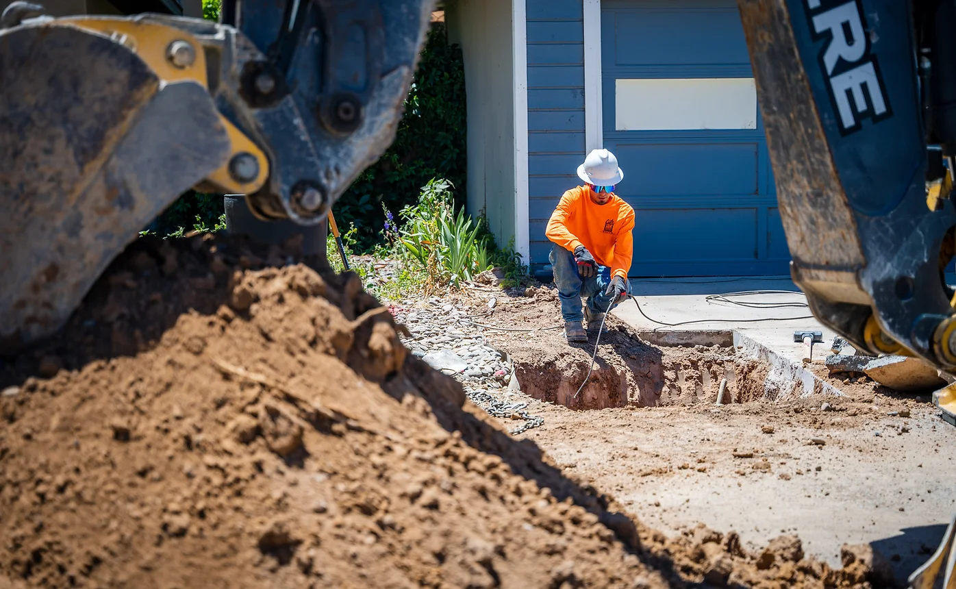 A construction worker in orange and an excavator digging near a house.