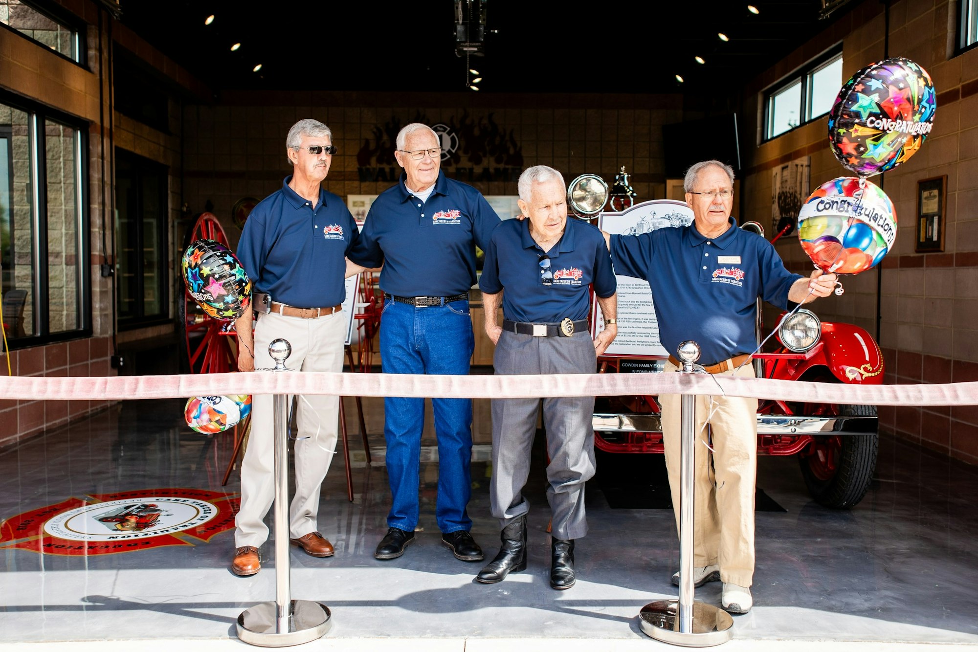 Four men in matching shirts at a ribbon-cutting ceremony with balloons and a vintage fire truck in the background.
