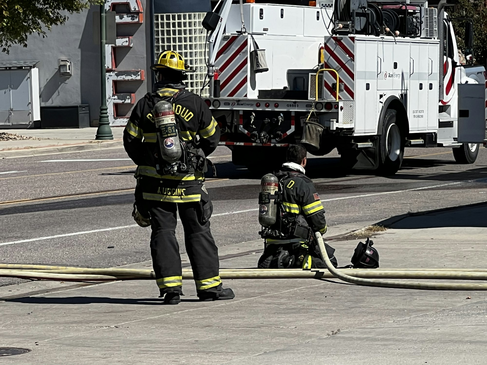 Firefighters with gear and hoses stand and kneel by a street