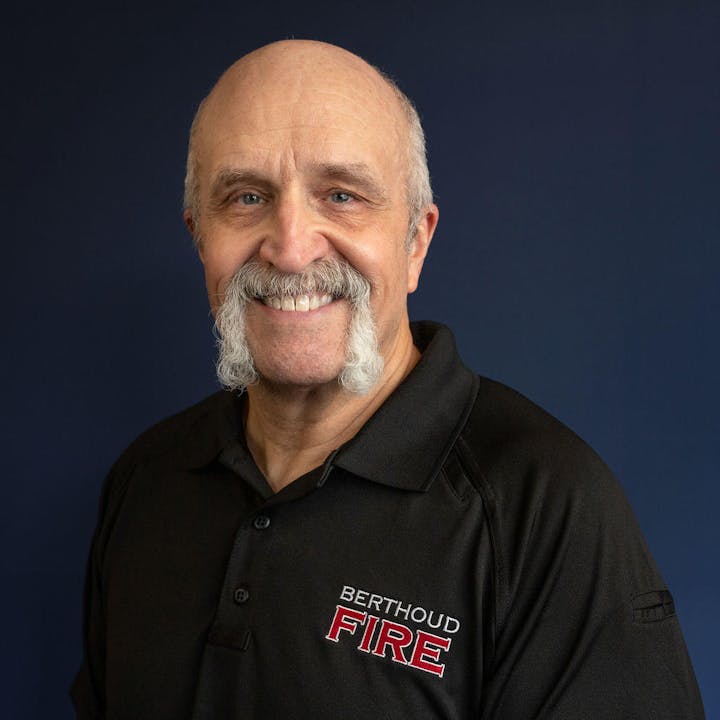 Smiling man with mustache wearing a "Berthoud Fire" shirt on a blue background.