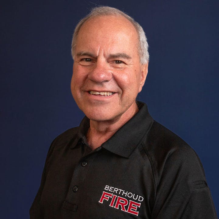 A smiling person wearing a shirt with "Berthoud FIRE" text, against a blue background.