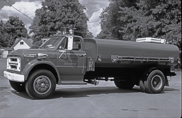 A vintage fire truck from a fire district, captured in black and white, parked among trees.