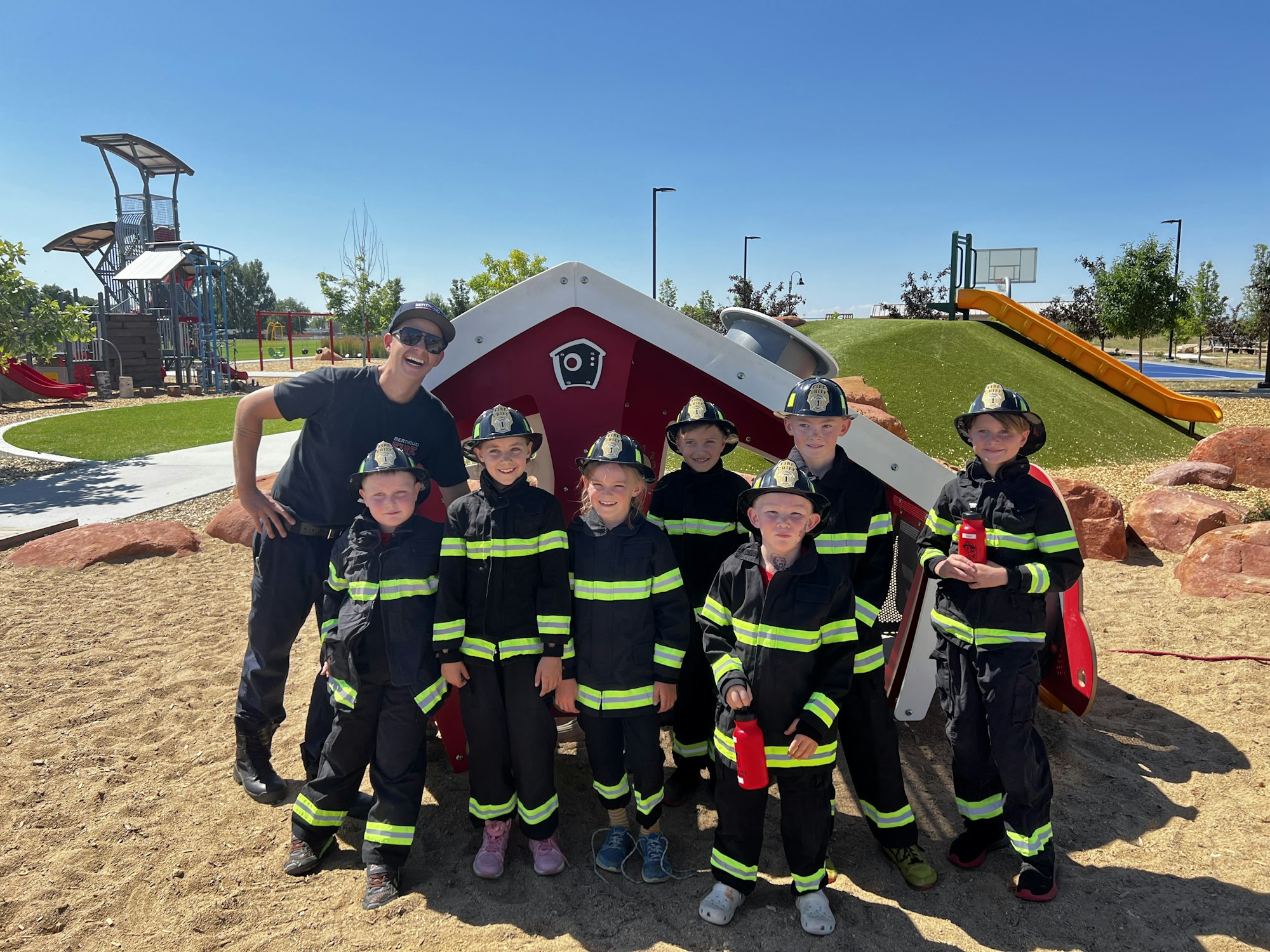 A group of kids in firefighter outfits with an adult, posing in a playground setting.