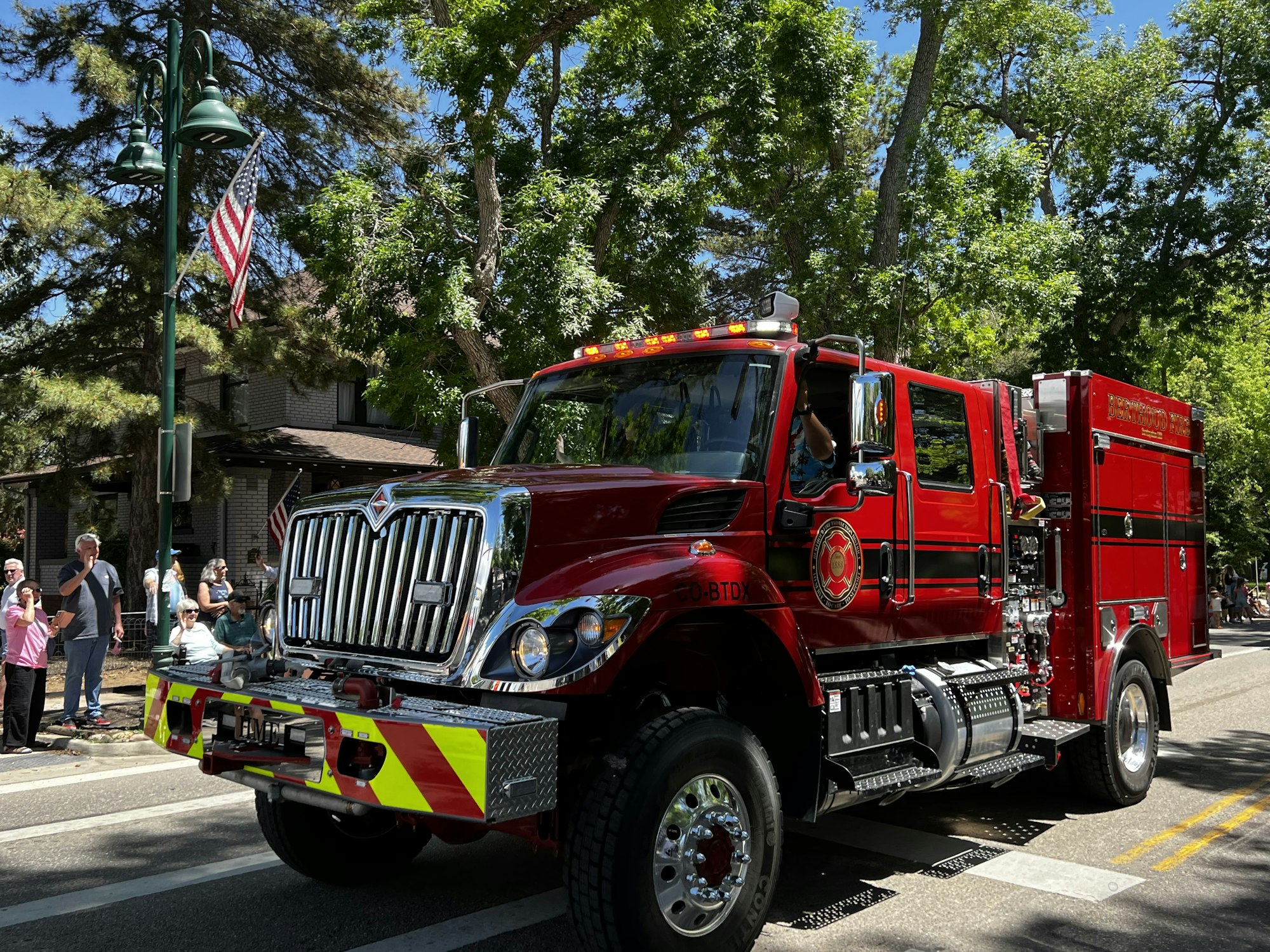 A red fire truck in a parade, surrounded by trees and onlookers, with an American flag nearby.