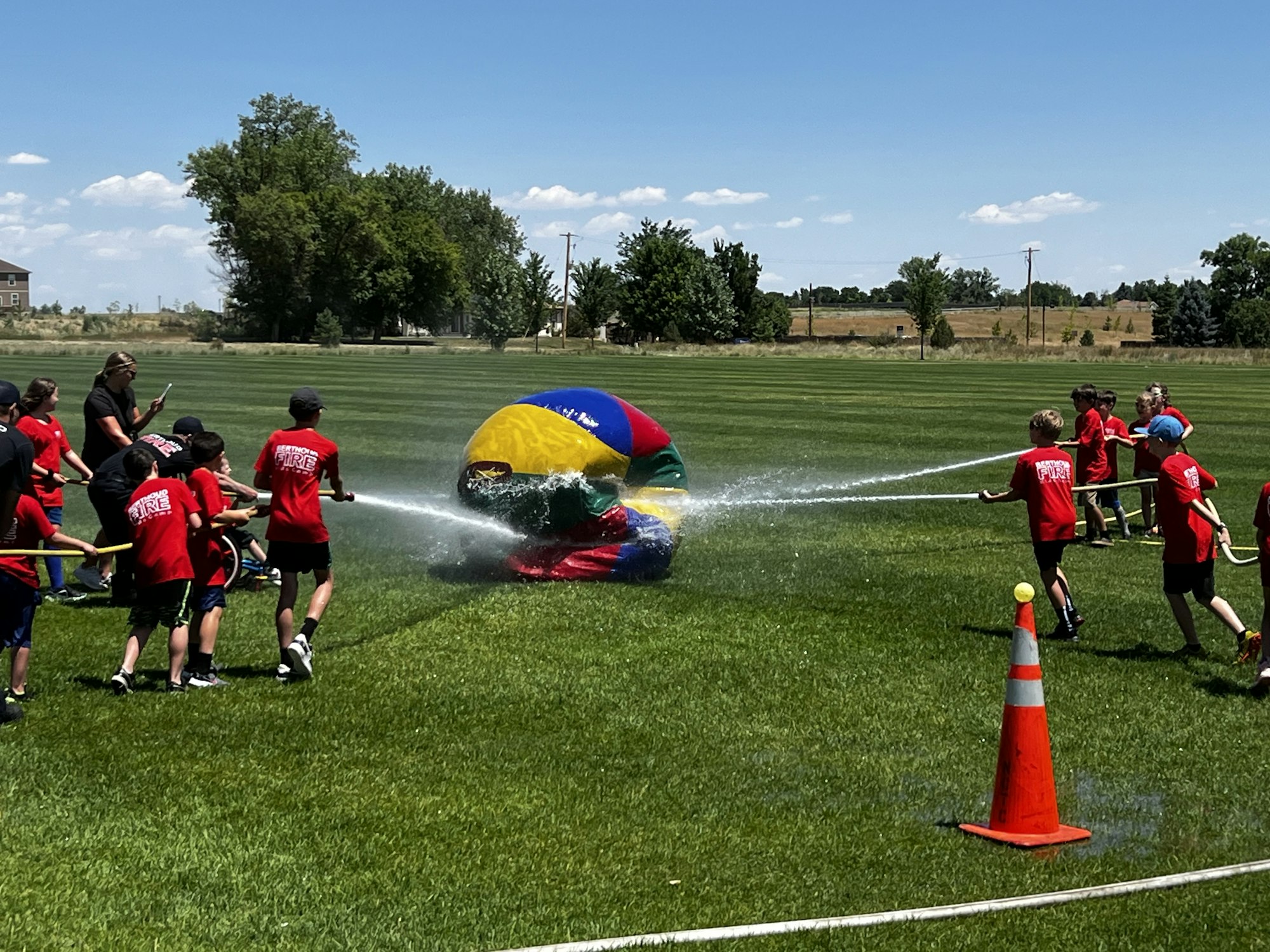 Kids in red shirts having fun with hoses spraying water on a large colorful ball in a grassy field.