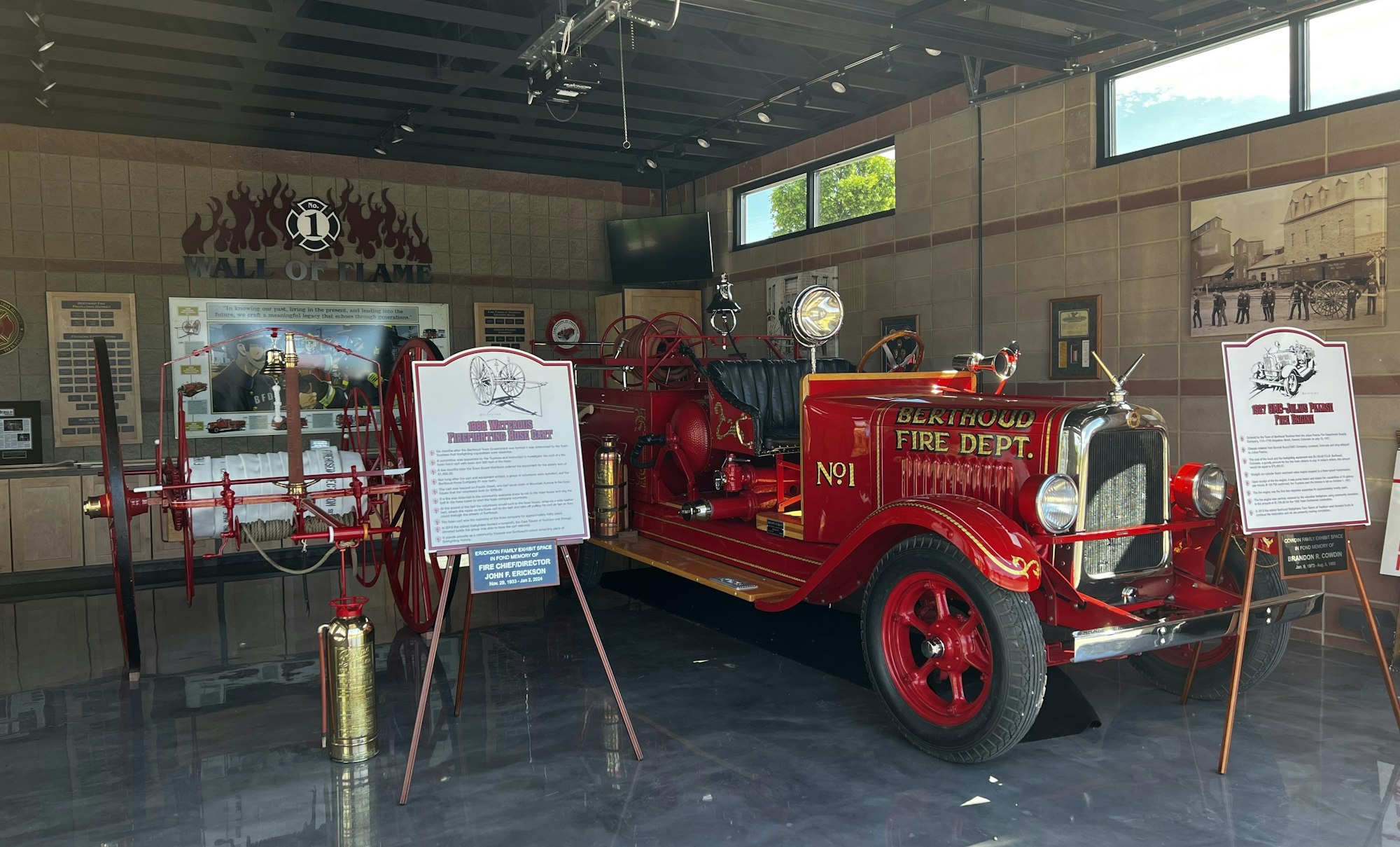 Vintage firetruck displayed in a museum with informational signs, "Wall of Flame" backdrop, and fire-fighting equipment.