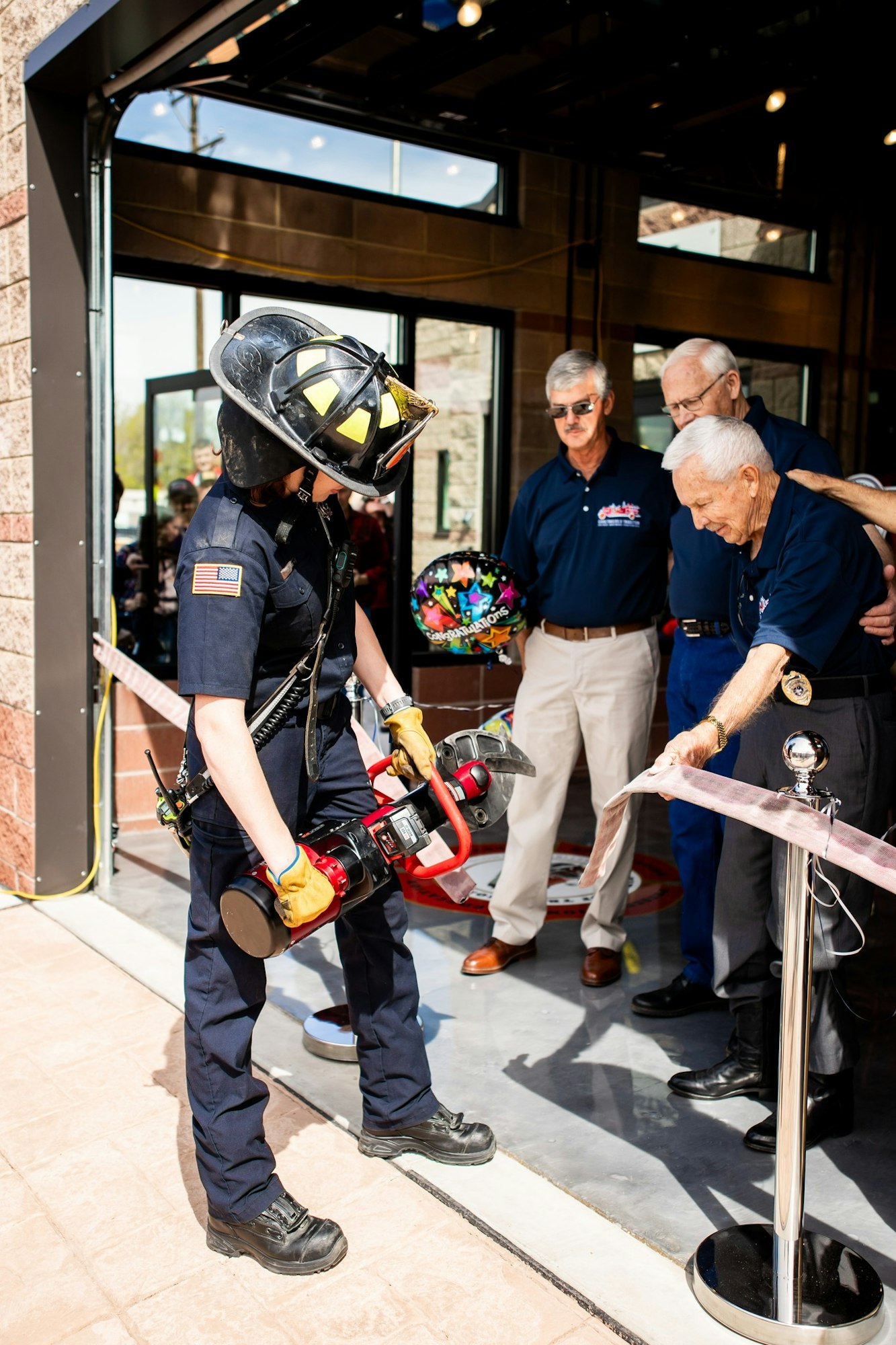 A firefighter with a tool, others watching a ribbon-cutting ceremony.