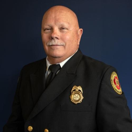 A portrait of a man with a moustache in a formal firefighter uniform with badge and gold buttons, against a blue background.