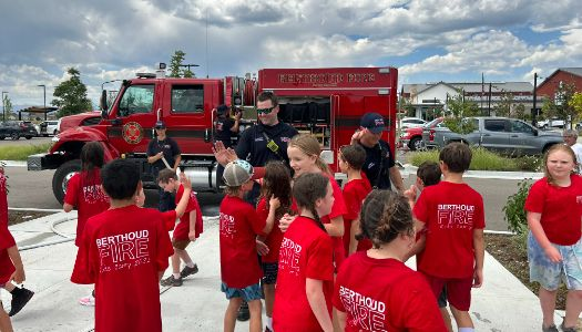 Children in red "Berthoud Fire" T-shirts interacting with firefighters near a fire engine.