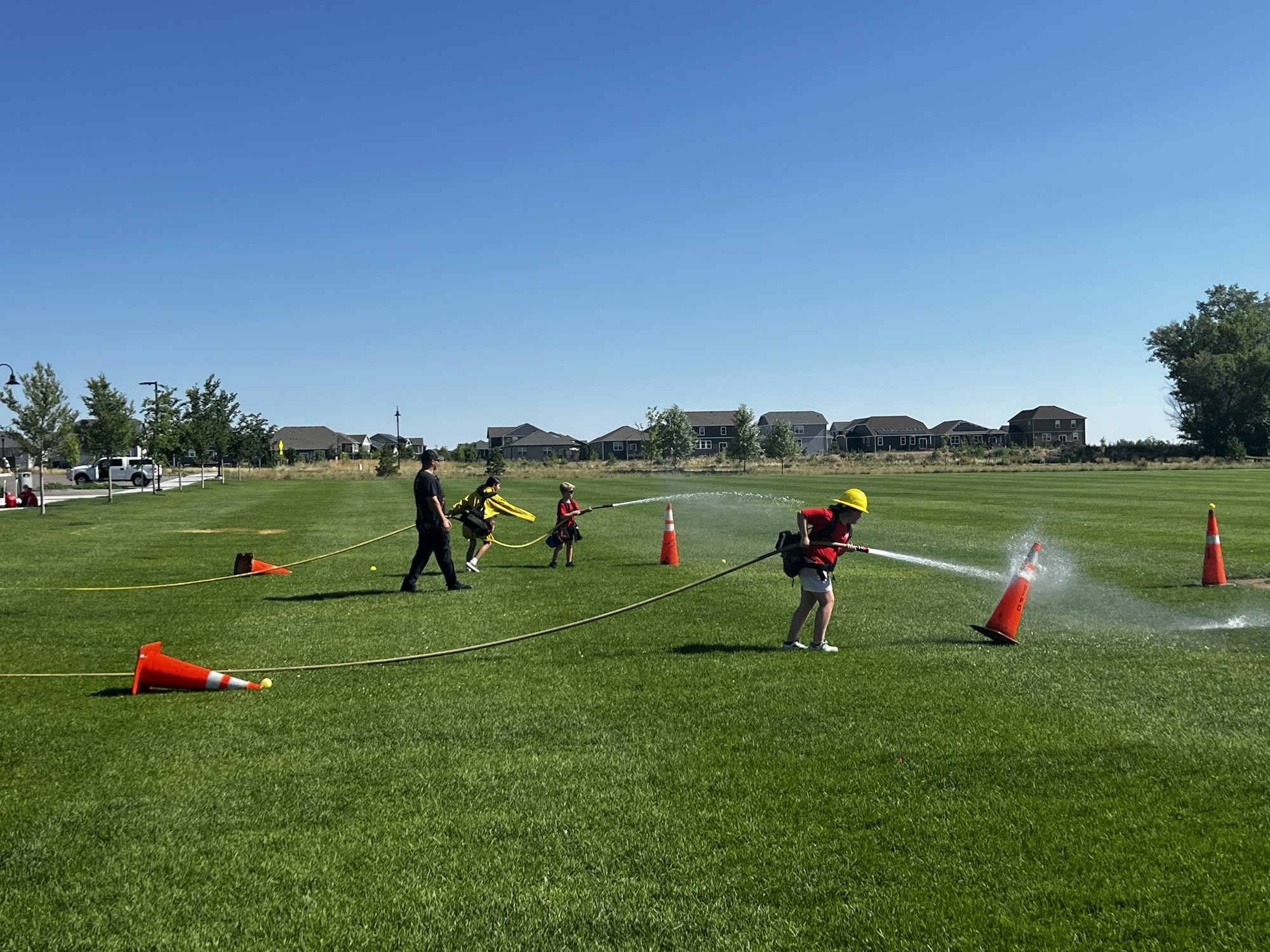 Children in a park spraying water from hoses at traffic cones, supervised by an adult.