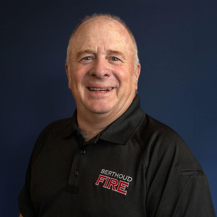 A smiling man in a black shirt with "Berthoud Fire" embroidered on it against a blue background.