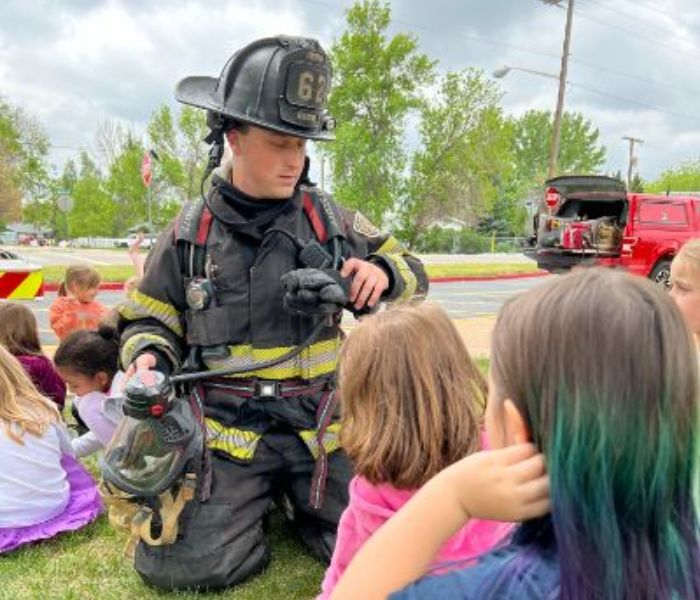 A firefighter is demonstrating equipment to children on grass with a fire truck in the background.
