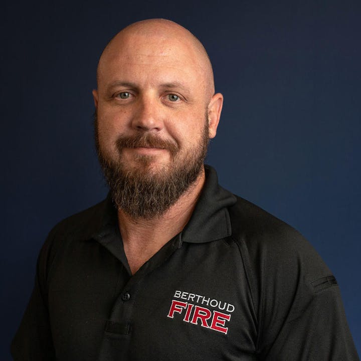 A bald man with a beard in a black shirt with "BERTHOUD FIRE" embroidered on it, blue background.