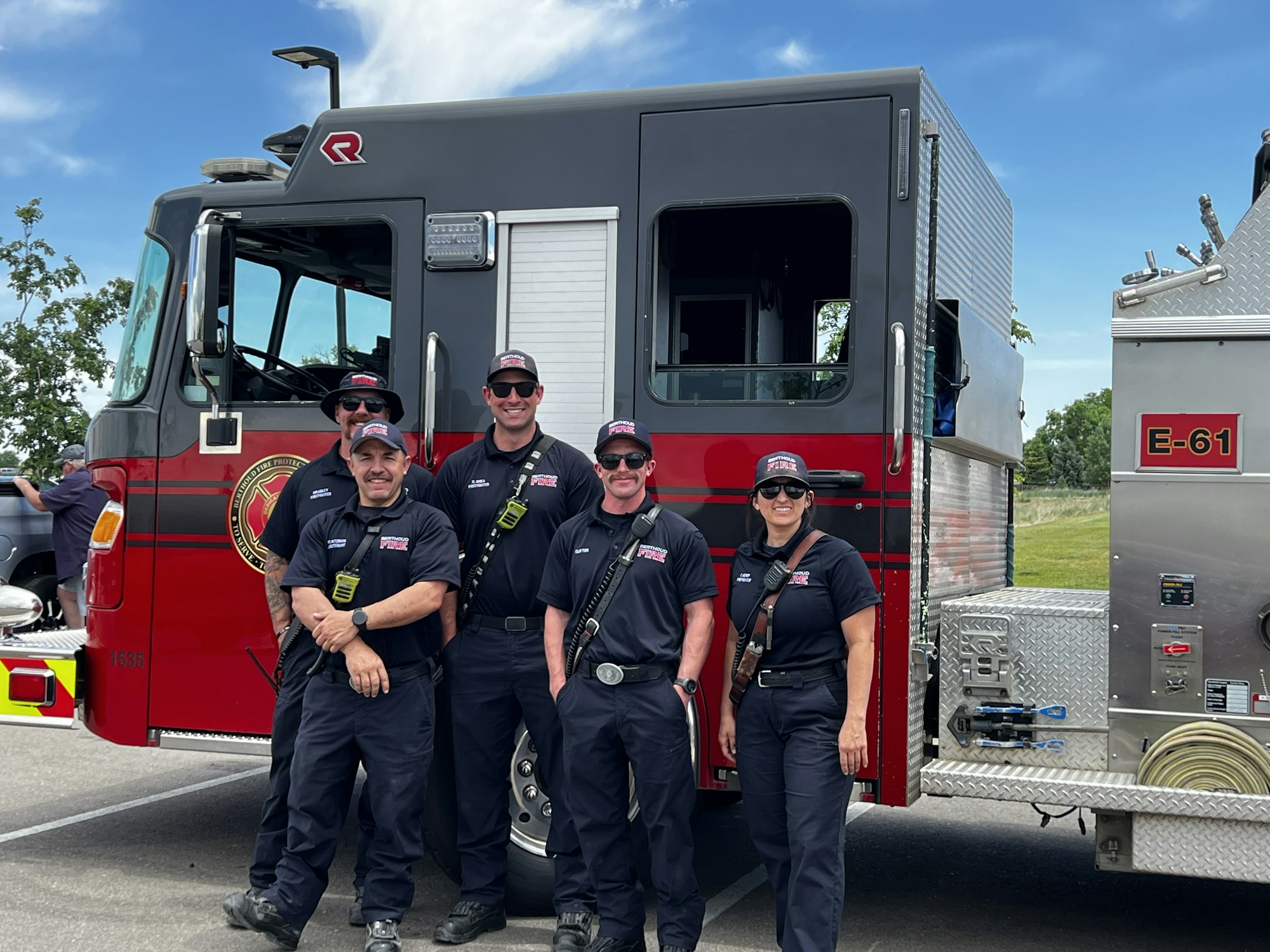 Five firefighters posing in front of a fire truck labeled "E-61" under a clear blue sky.