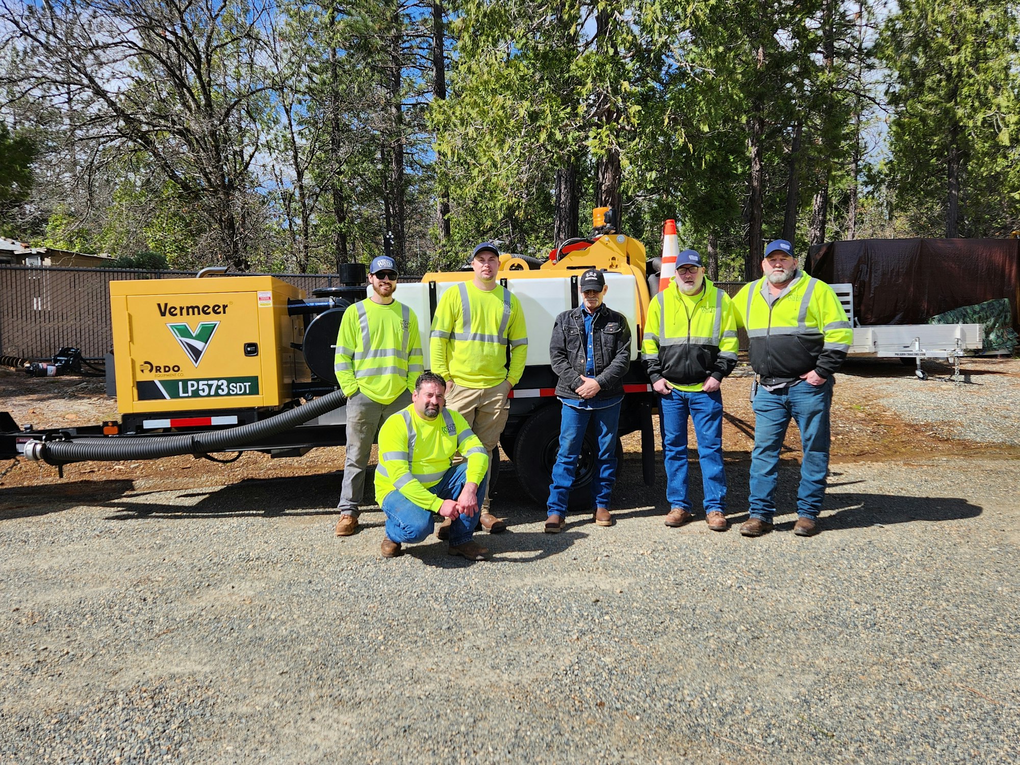 A group of workers in high-visibility vests posing in front of industrial equipment outdoors.
